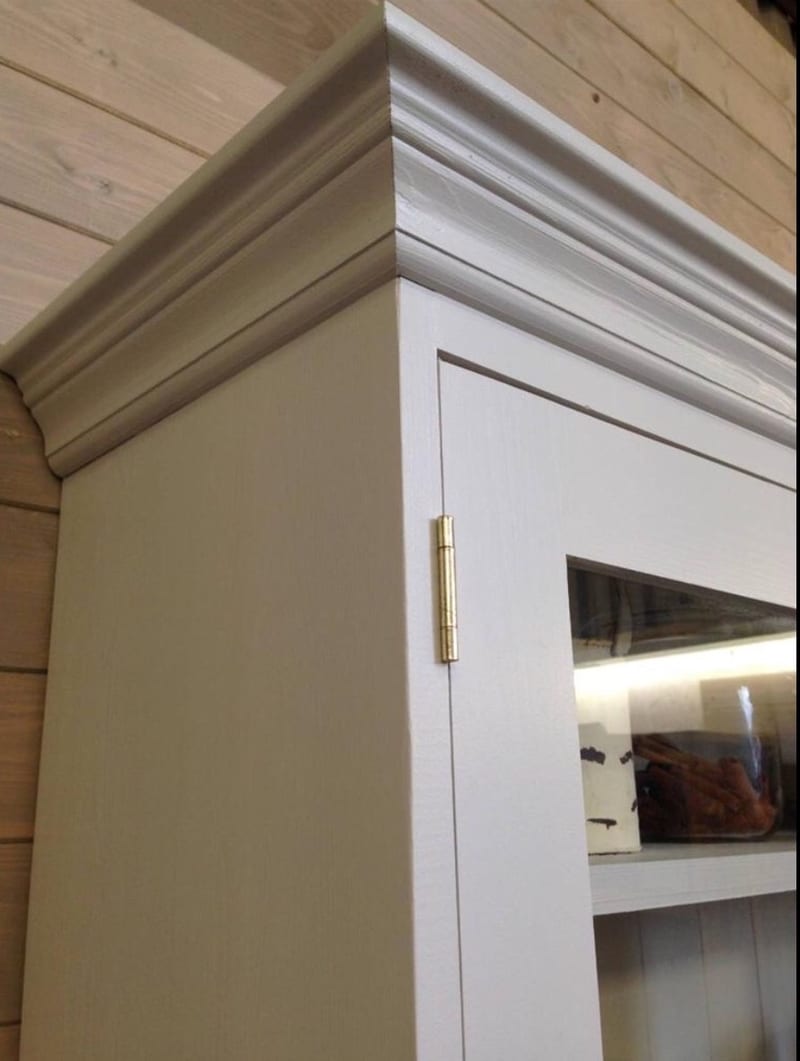 Close view of the top corner of the dresser where the moulded cornice meets the painted side panel, with a brass hinge on the glazed door and pine plank walls behind