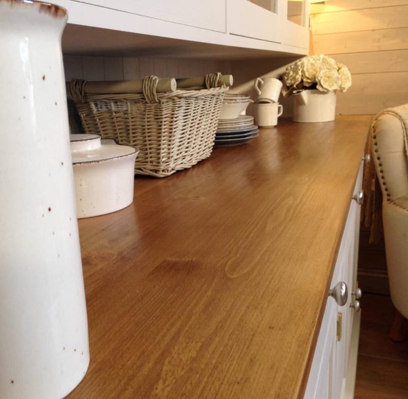 Close view along the waxed pine worktop showing the grain and sheen, with a stoneware jug, lidded pot, wicker baskets, stacked plates and cream jugs set out towards the far end