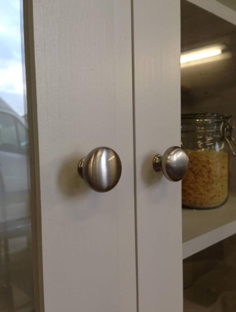 Extreme close-up of two brushed nickel knobs on the glazed doors, showing their turned shape and the painted door stiles, with a lit shelf and a jar of pasta behind the glass
