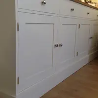 Low angled view of the painted base cupboards from the end, showing four panelled doors, brass butt hinges, brushed nickel knobs and the deep plinth against pale wood flooring