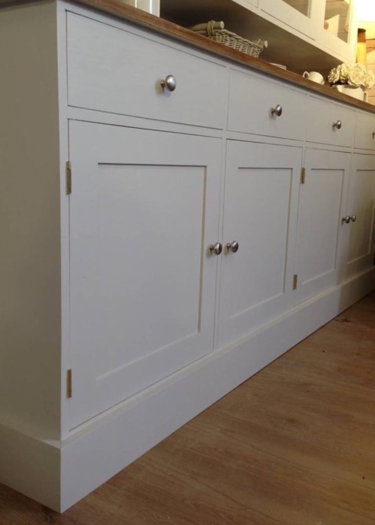 Low angled view of the painted base cupboards from the end, showing four panelled doors, brass butt hinges, brushed nickel knobs and the deep plinth against pale wood flooring
