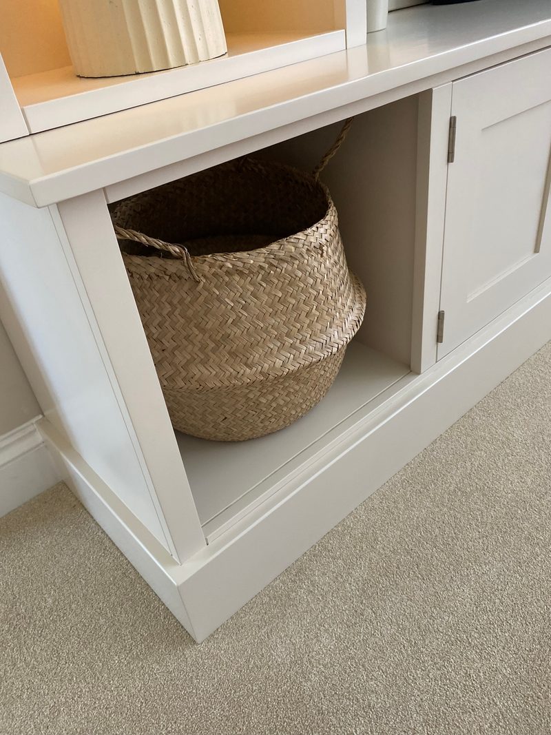 Close view of the lower corner of the painted media unit showing an open cubby with a woven seagrass basket, a shaker cupboard door on brushed metal hinges and the plinth on cream carpet