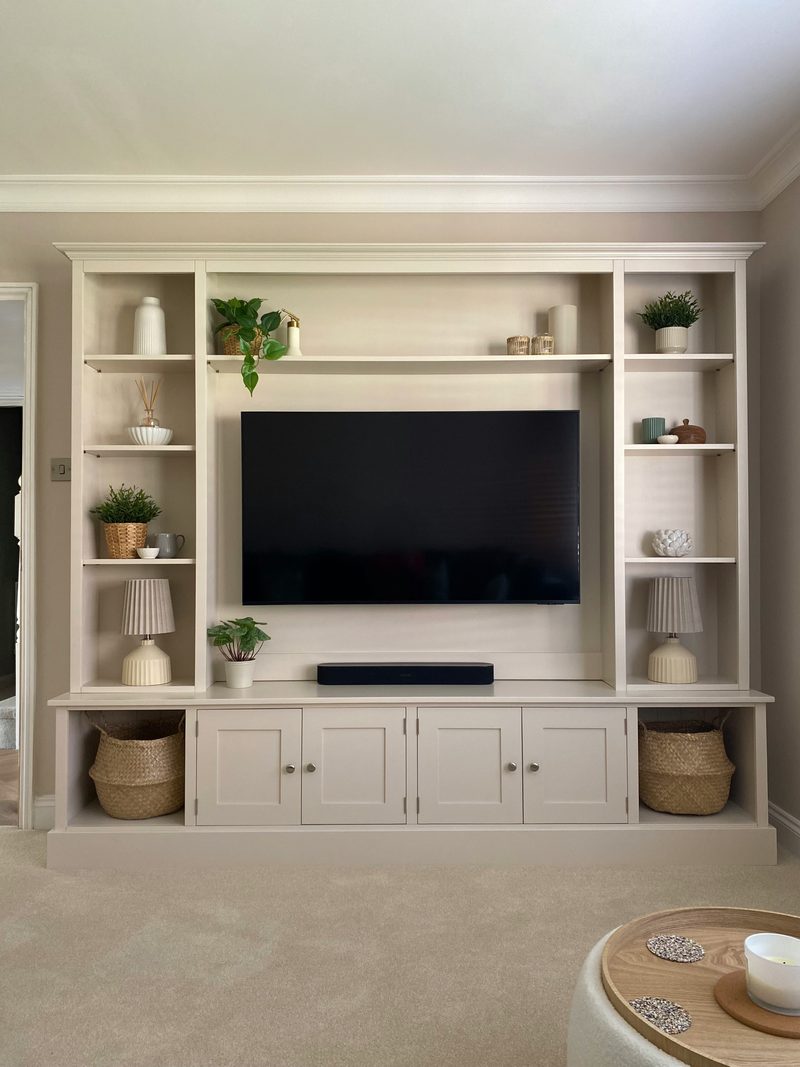 Straight-on view of the taupe painted media wall with open shelving to each side, the television and soundbar on the central panel, four cupboard doors below and baskets in the end cubbies
