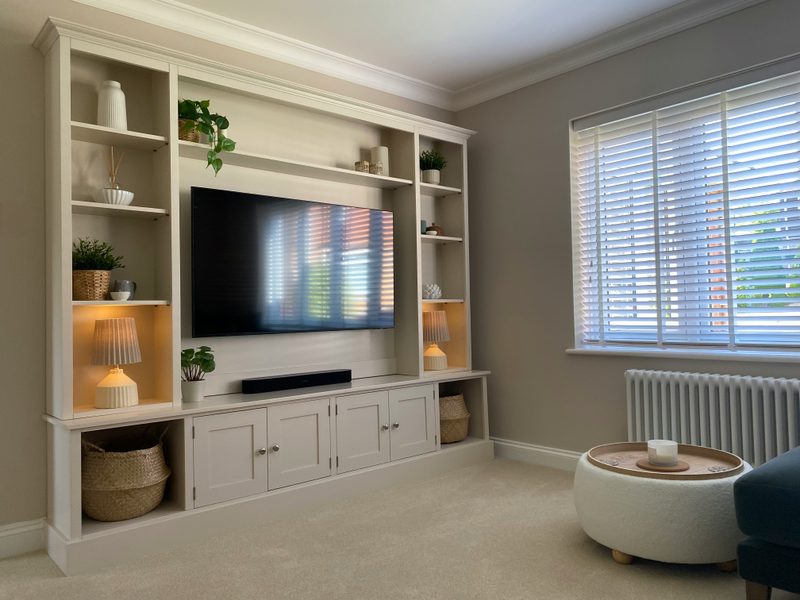 Angled room view of the media wall with the television switched on and shelf lamps lit, potted plants, baskets in the end cubbies and a round footstool beside a blind-covered window