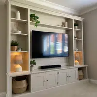 Three-quarter view of the media wall showing the stepped cornice, lamps lit on the open shelves of both towers, the television above the soundbar and four shaker doors with brushed nickel knobs