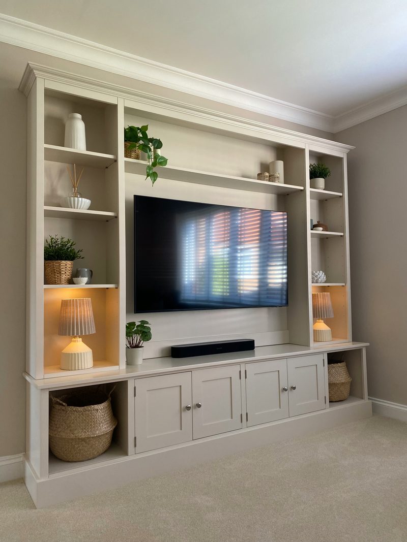 Three-quarter view of the media wall showing the stepped cornice, lamps lit on the open shelves of both towers, the television above the soundbar and four shaker doors with brushed nickel knobs