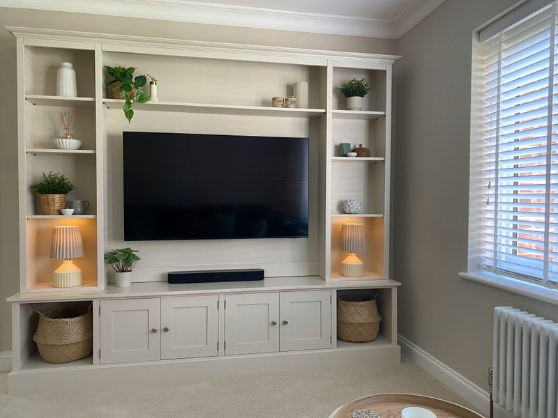 Wide room view of the media wall with lit shelf lamps, a blank television screen, seagrass baskets in the end cubbies, a column radiator and a window with white venetian blinds alongside
