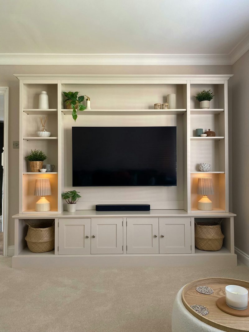 Front view of the media wall with both shelf lamps lit and the television off, showing the long display shelf of pots, plants and candles, the soundbar and four cupboards on the plinth