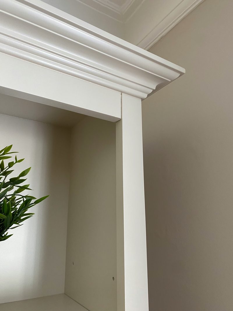 Close view of the top corner of the media wall showing the layered moulded cornice, the painted upright and shelf edge, an artificial plant on the shelf and the coving on the wall behind