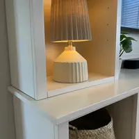 Detail of a ribbed cream table lamp lit on a shelf within the painted tower, showing the shelf pin holes, the worktop edge below, a seagrass basket beneath and a window blind to the right