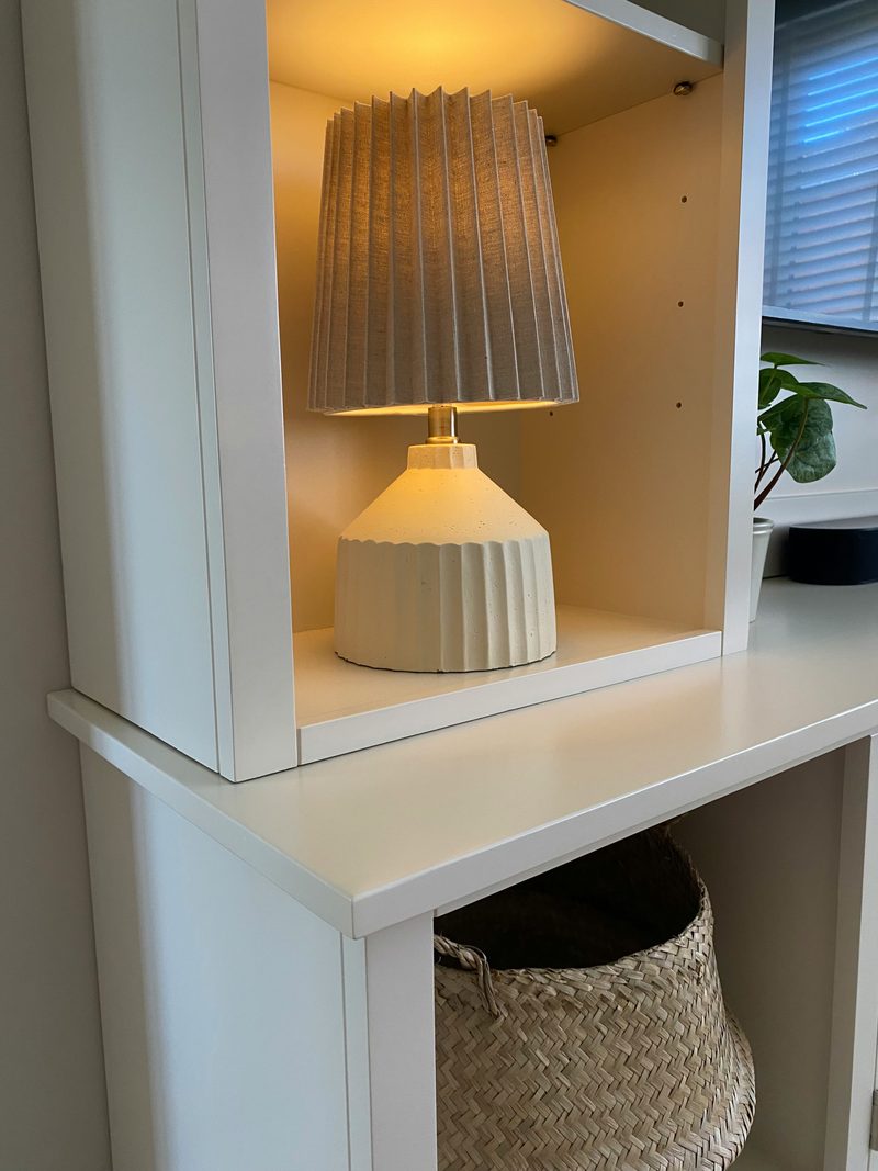 Detail of a ribbed cream table lamp lit on a shelf within the painted tower, showing the shelf pin holes, the worktop edge below, a seagrass basket beneath and a window blind to the right