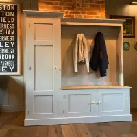 Pale grey boot room unit with a tall cupboard beside a coat hook rail and an oak topped bench over two cupboards, against a panelled wall below an exposed brick chimney breast