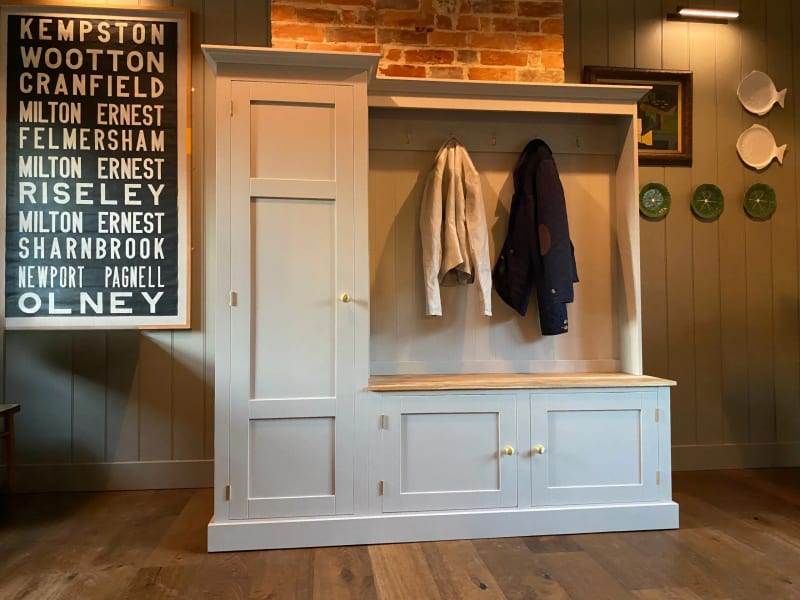 Pale grey boot room unit with a tall cupboard beside a coat hook rail and an oak topped bench over two cupboards, against a panelled wall below an exposed brick chimney breast