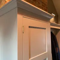 Close view of the top corner of the pale grey painted boot room unit, showing the moulded cornice, a brass butt hinge on the tall shaker door and an exposed brick chimney breast behind