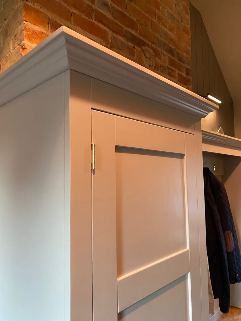 Close view of the top corner of the pale grey painted boot room unit, showing the moulded cornice, a brass butt hinge on the tall shaker door and an exposed brick chimney breast behind