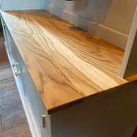 Close view along the timber bench top of the boot room unit, showing the streaked grain and figuring of the oiled surface, the painted back rail and brass knobs and hinges on the cupboards below