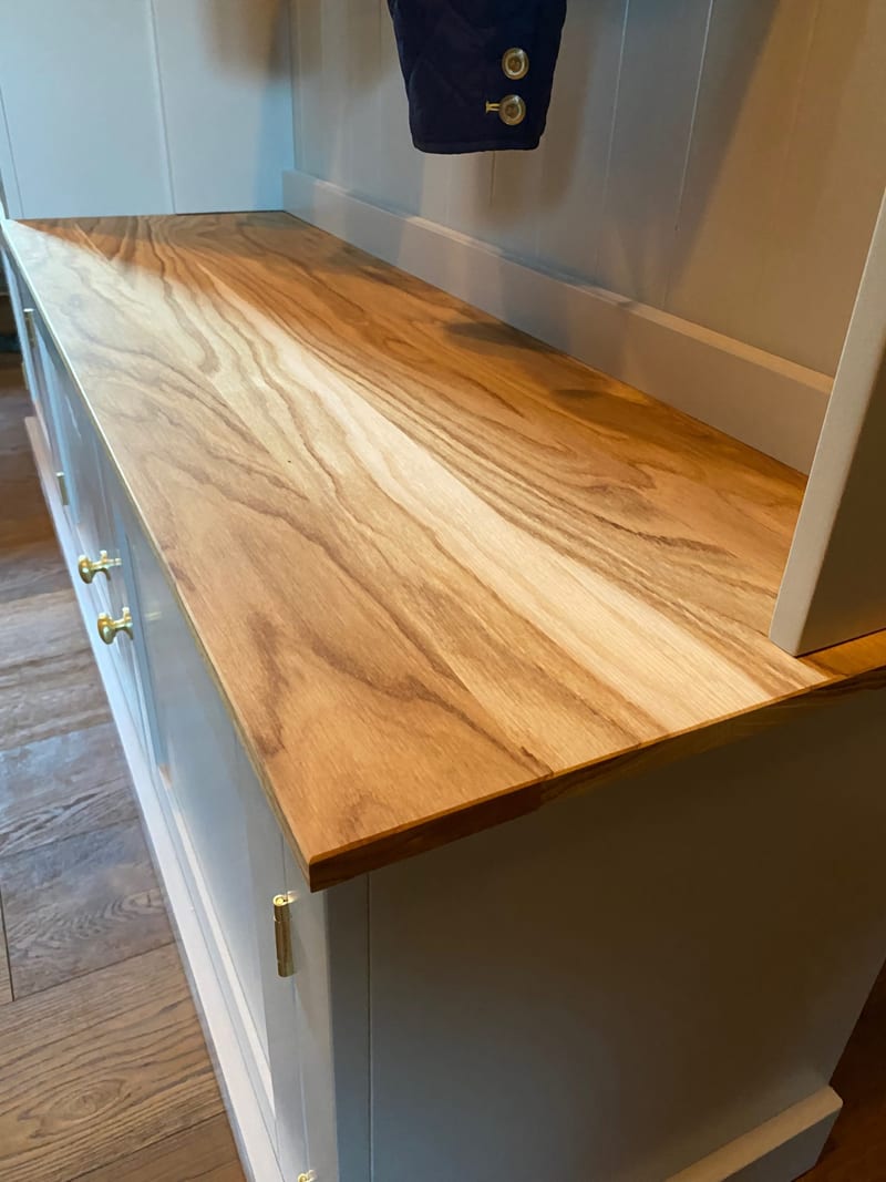 Close view along the timber bench top of the boot room unit, showing the streaked grain and figuring of the oiled surface, the painted back rail and brass knobs and hinges on the cupboards below
