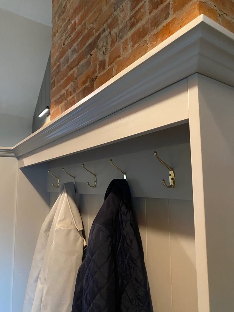 Detail of the cornice above the coat rail, showing a row of antique brass double hooks on the tongue and groove backboard with a cream jacket and a navy quilted coat hanging