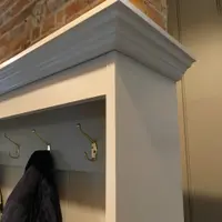 Close view of the outer corner of the cornice above the coat rail, showing the moulded profile, the painted end panel, three brass hooks and a navy quilted coat against the brick chimney