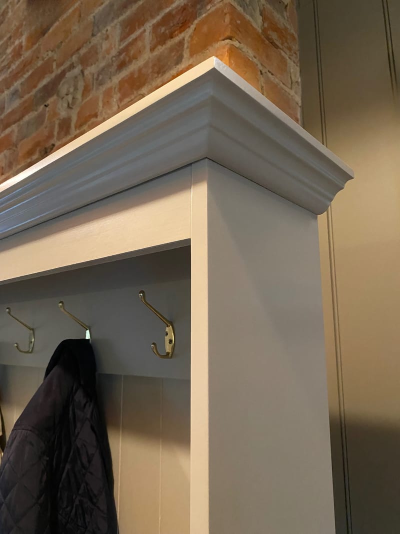 Close view of the outer corner of the cornice above the coat rail, showing the moulded profile, the painted end panel, three brass hooks and a navy quilted coat against the brick chimney