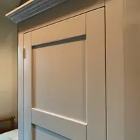 Close view of the tall cupboard door on the boot room unit, showing the shaker frame, the moulded cornice above, a brass hinge and the painted end panel against a green panelled wall