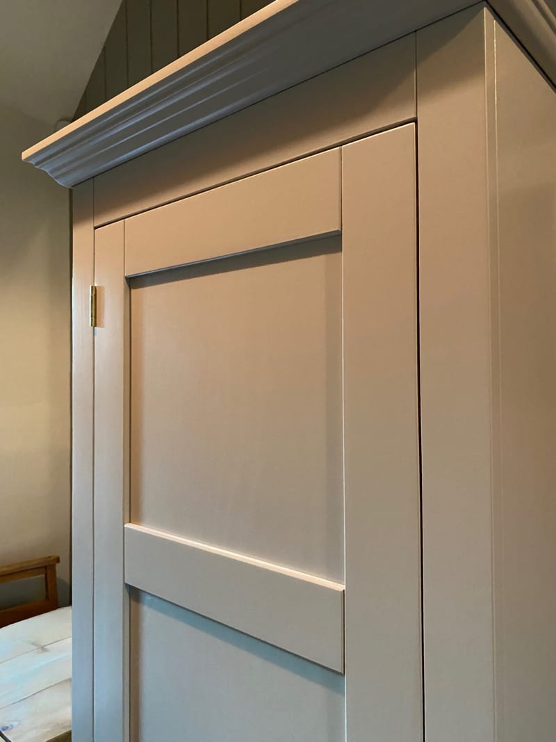 Close view of the tall cupboard door on the boot room unit, showing the shaker frame, the moulded cornice above, a brass hinge and the painted end panel against a green panelled wall