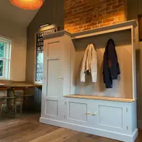 Wide room view of the boot room unit against a green panelled wall and brick chimney breast, with two coats on the brass hooks, a wicker pendant light, a bus blind print and plank flooring