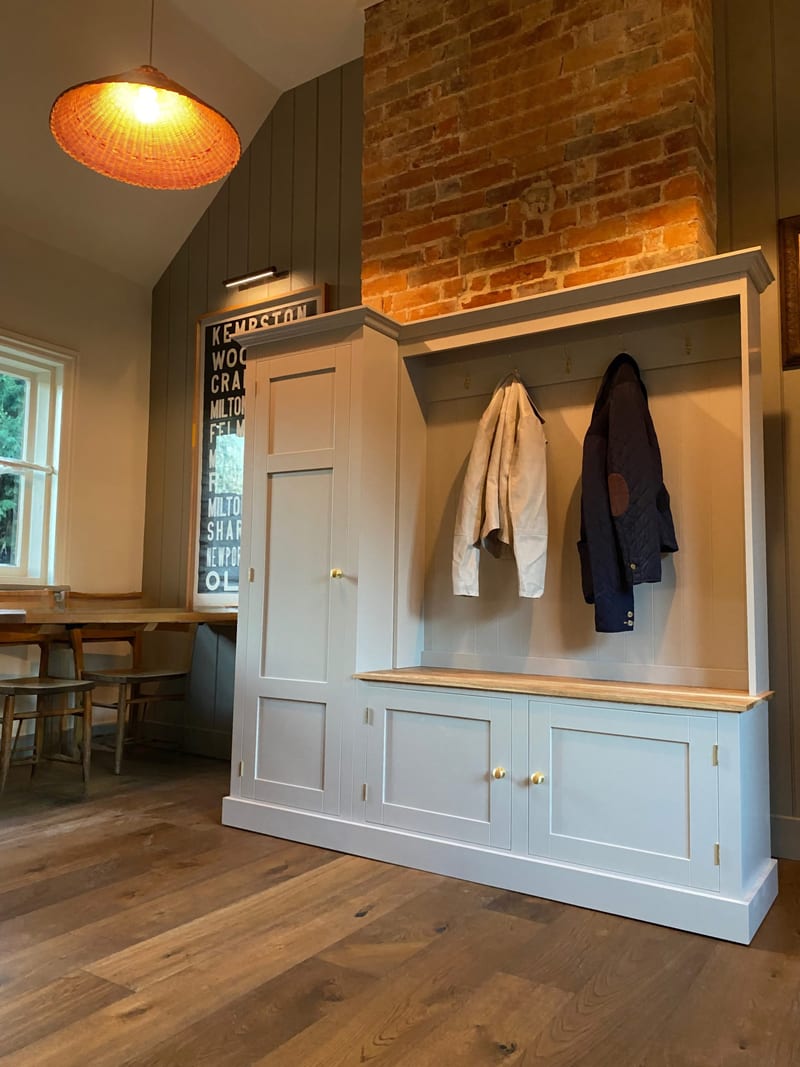 Wide room view of the boot room unit against a green panelled wall and brick chimney breast, with two coats on the brass hooks, a wicker pendant light, a bus blind print and plank flooring