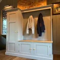 Angled view of the boot room unit showing the tall cupboard, the open coat rail with brass hooks, the timber bench top and two panelled cupboard doors with brass knobs above the plinth