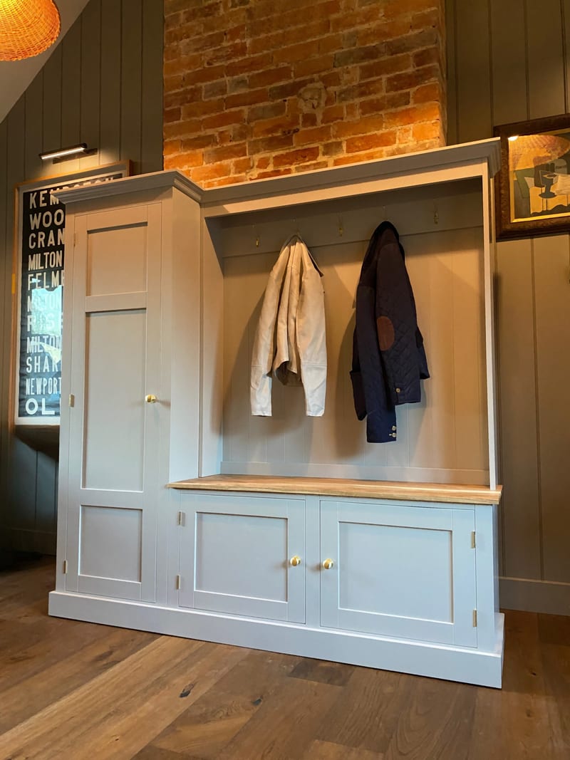 Angled view of the boot room unit showing the tall cupboard, the open coat rail with brass hooks, the timber bench top and two panelled cupboard doors with brass knobs above the plinth
