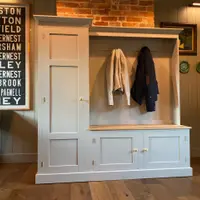 Front view of the boot room unit with the tall single cupboard to the left, a coat rail holding a cream jacket and navy coat, a timber bench top and two brass-knobbed cupboards below