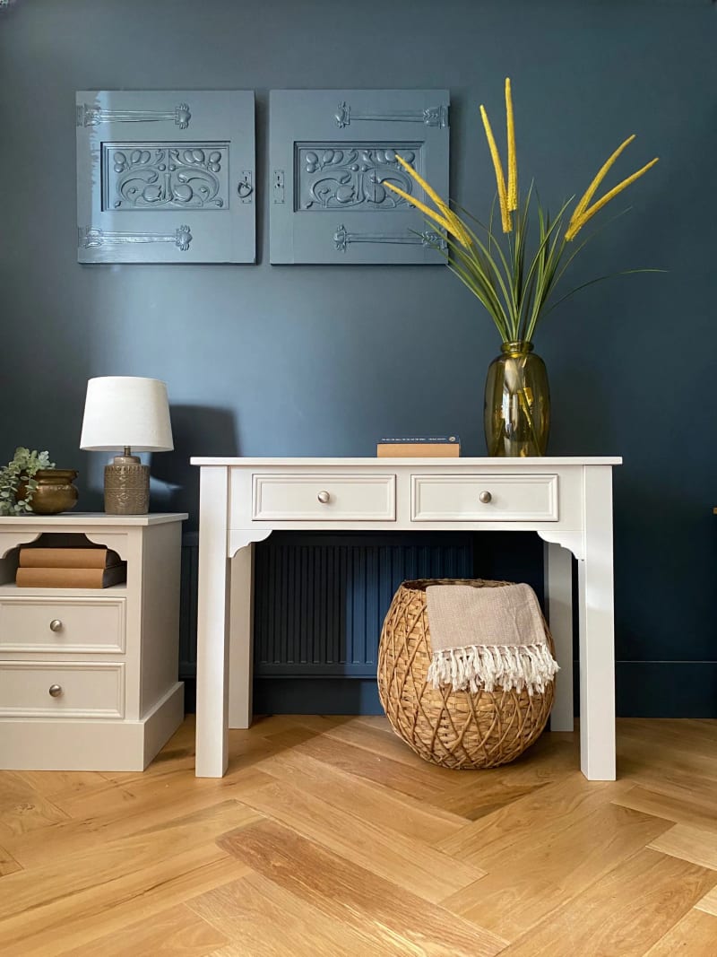 Cream painted Carlton console table with two drawers and shaped corner brackets, against a deep blue wall with a woven basket beneath and a bedside cabinet alongside