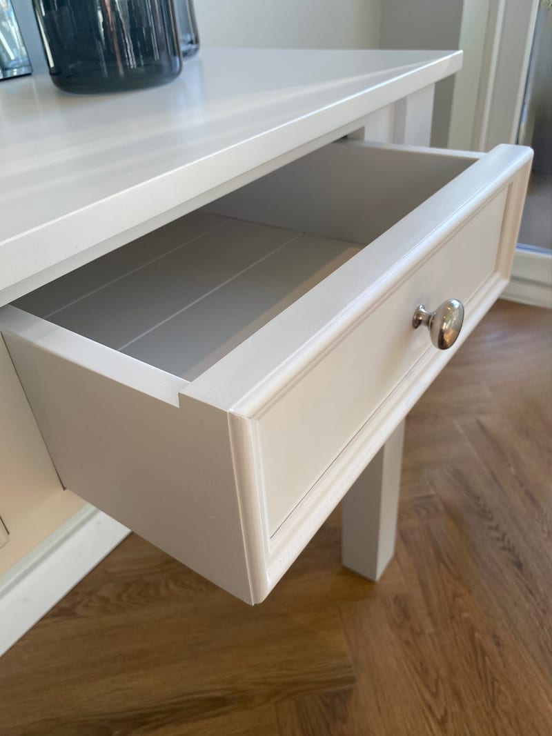Close view of one drawer pulled open on the white painted console table, showing the moulded drawer front, round chrome knob and grey painted interior with a boarded base