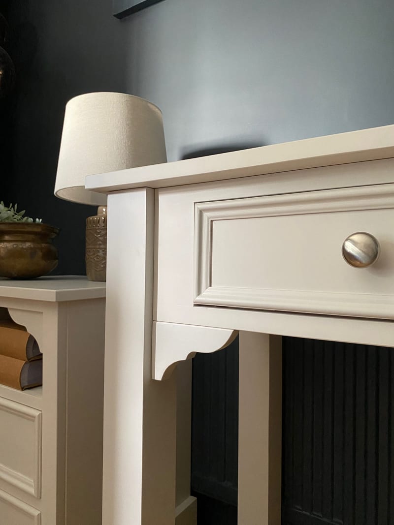Corner detail of the cream painted console table showing the moulded drawer front, brushed nickel knob and shaped bracket at the leg, against a dark blue wall beside a lamp and brass pot