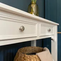 Angled close view of both drawer fronts and the top edge of the cream painted console table, with brushed nickel knobs, an olive glass vase above and a woven basket with a fringed throw below