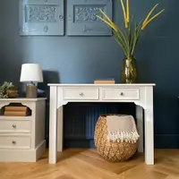 Full front view of the cream painted console table with two drawers and round knobs, set against a deep blue wall with framed relief panels above and a woven basket underneath