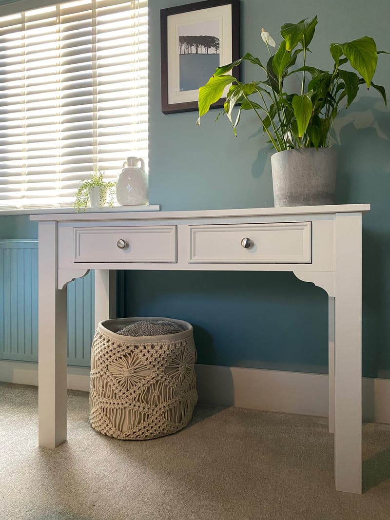 Full view of the white painted console table with two drawers standing below a window with wooden slat blinds, a peace lily on top and a macrame basket beneath on beige carpet