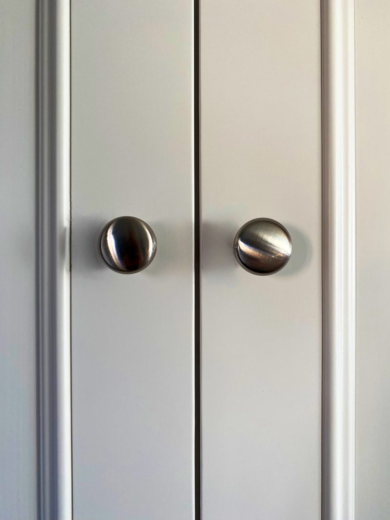 Close-up of the two brushed nickel knobs where the wardrobe doors meet, showing the pale painted timber, the fine gap between the doors and the moulded edge of the side frame