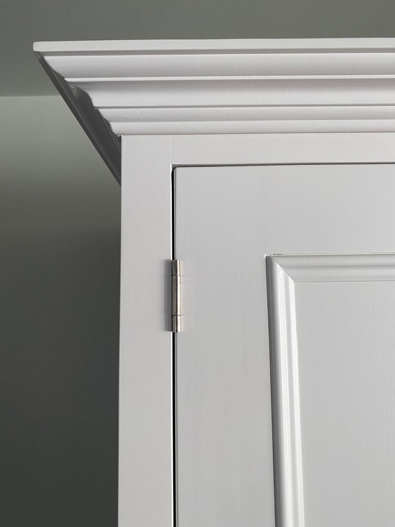 Detail of the top corner of the white painted wardrobe, showing the stepped moulded cornice, a brushed nickel butt hinge and the raised panel edge of the door