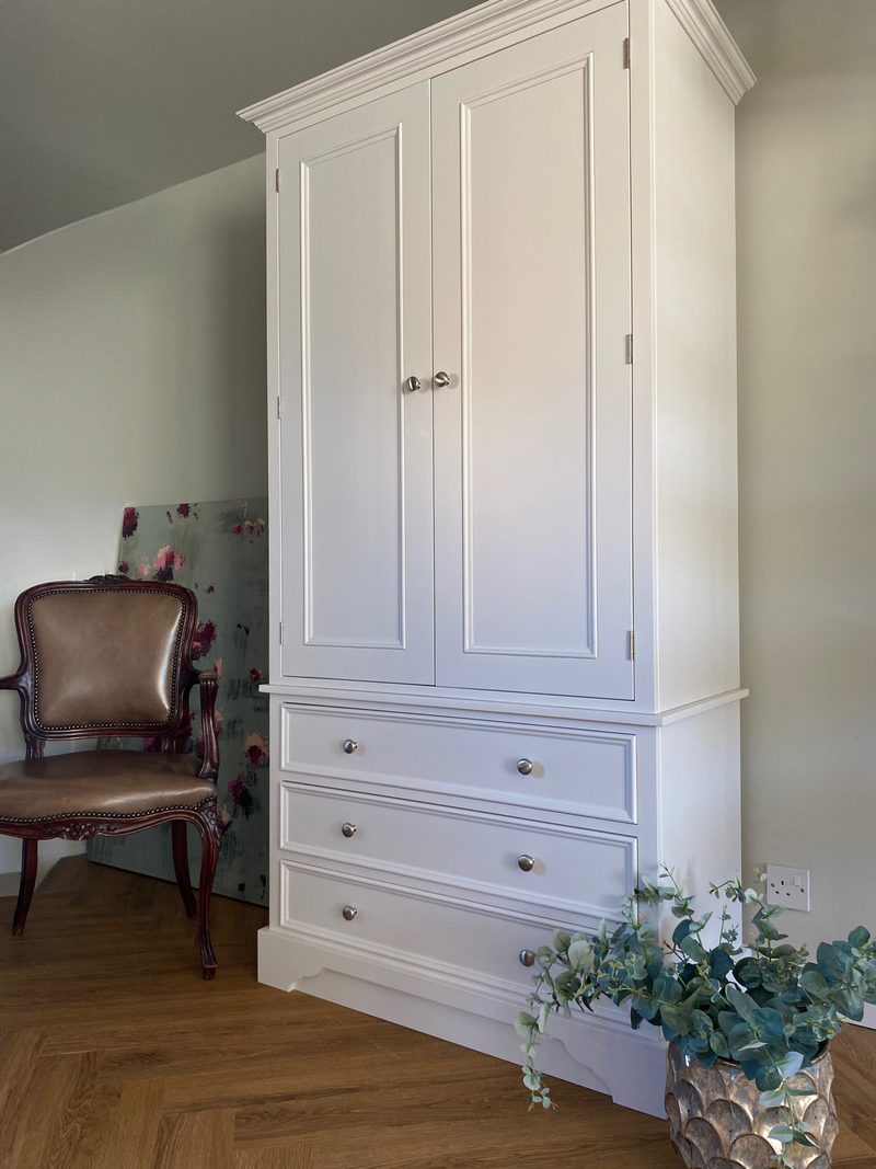 Angled full view of the white painted wardrobe with two panelled doors above three deep drawers, all with round nickel knobs, beside a leather armchair and eucalyptus on herringbone oak flooring