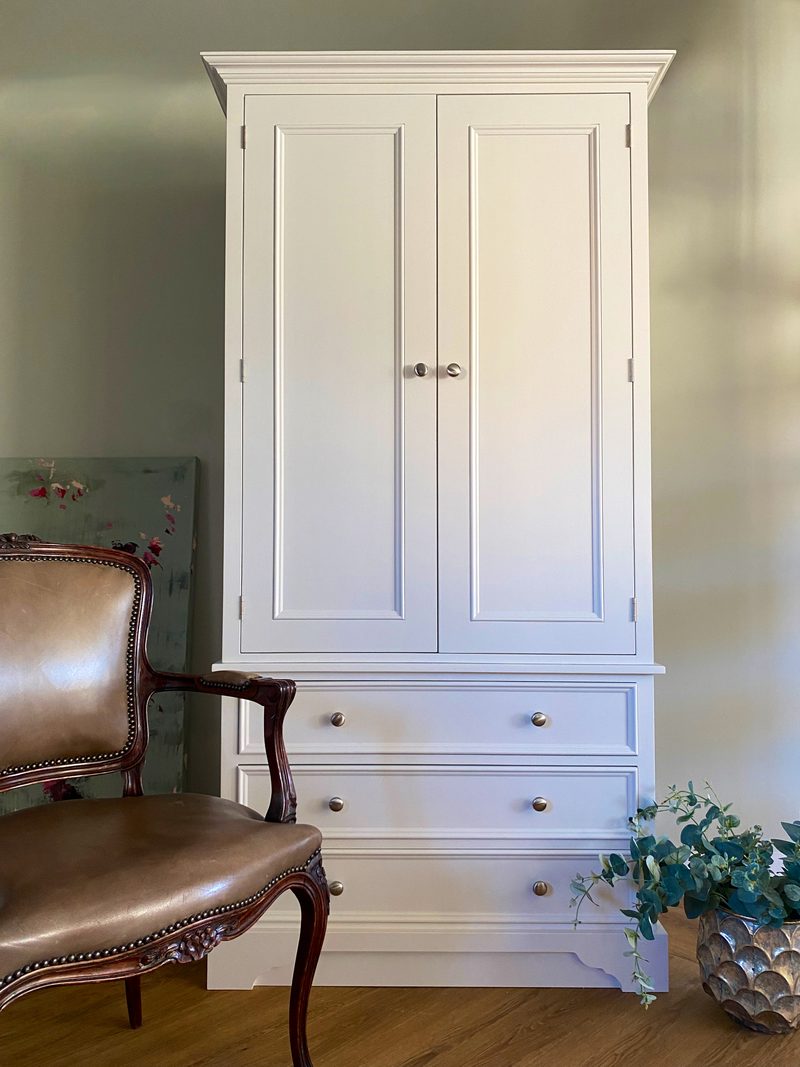 Straight-on view of the painted wardrobe showing the moulded cornice, two long panelled doors, three full-width drawers and the shaped plinth, with a leather framed armchair in front