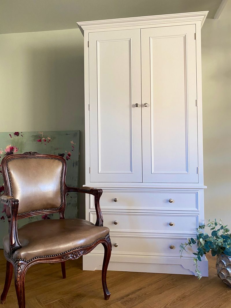 Side-angled view of the tall painted wardrobe against a sage green wall, with the cornice, panelled doors and three drawers visible past an antique leather armchair on oak flooring