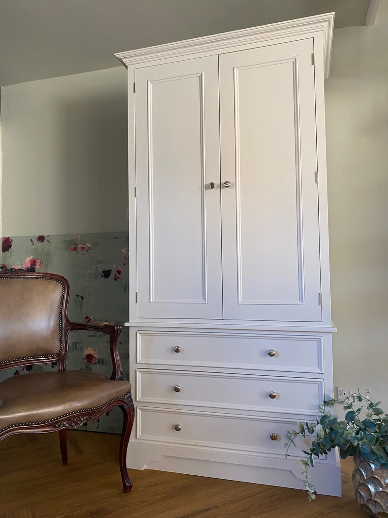Three-quarter view of the wardrobe showing the depth of the carcass and the projecting cornice, its two panelled doors closed on brushed nickel knobs above three drawers on a wood floor