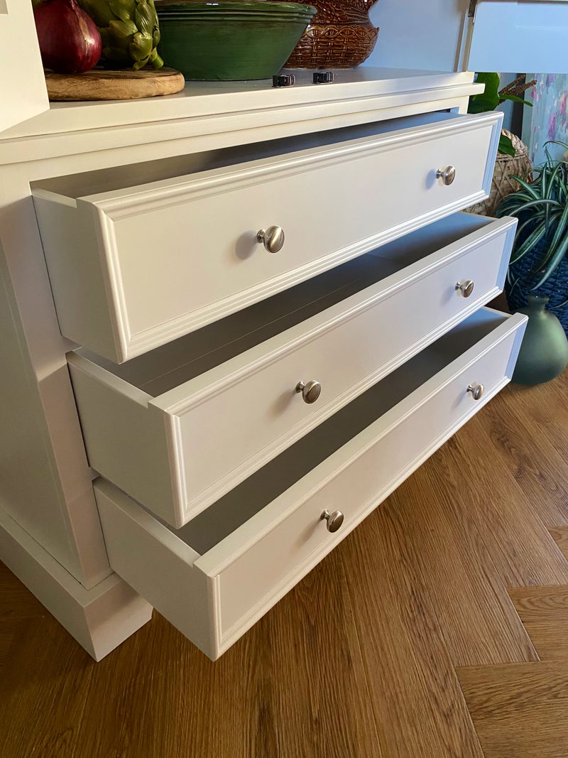 Detail of the three drawers pulled open on the base of a white painted kitchen dresser, showing the moulded fronts, round nickel knobs and painted drawer sides above a herringbone wood floor