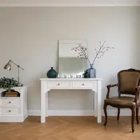 Wide view of the white painted dressing table in a pale grey room, dressed with a rectangular mirror, blossom branches and small bowls, with a matching bedside cabinet and antique chair alongside