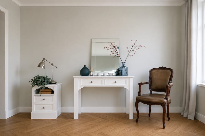 Wide view of the white painted dressing table in a pale grey room, dressed with a rectangular mirror, blossom branches and small bowls, with a matching bedside cabinet and antique chair alongside