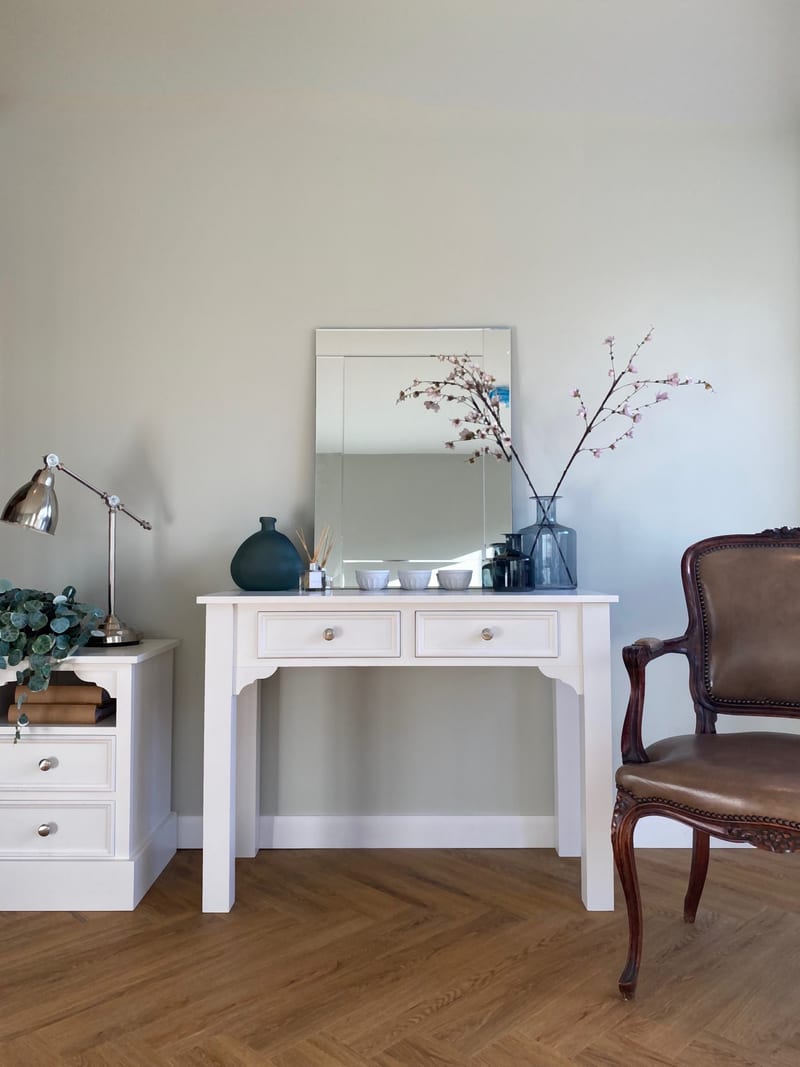 Portrait view of the dressing table styled with a bevelled mirror, dark ceramic vase, reed diffuser and three small white bowls, beside a bedside cabinet and a leather armchair