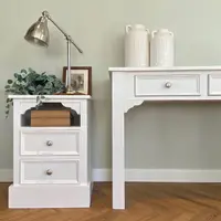 Close view of a small painted bedside cabinet with an open shelf and two drawers beside the end of the dressing table, with a chrome desk lamp, eucalyptus and a pair of textured white vases