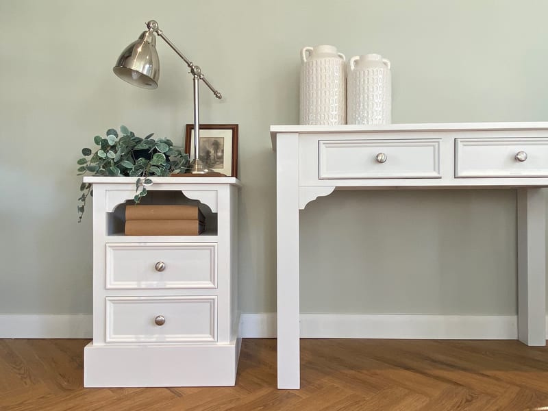 Close view of a small painted bedside cabinet with an open shelf and two drawers beside the end of the dressing table, with a chrome desk lamp, eucalyptus and a pair of textured white vases