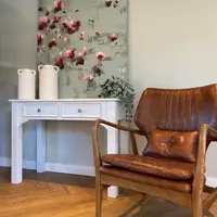 Full view of the white painted dressing table holding two textured ceramic vases below a leaning floral canvas, with a tan leather and oak armchair in front on herringbone wood flooring